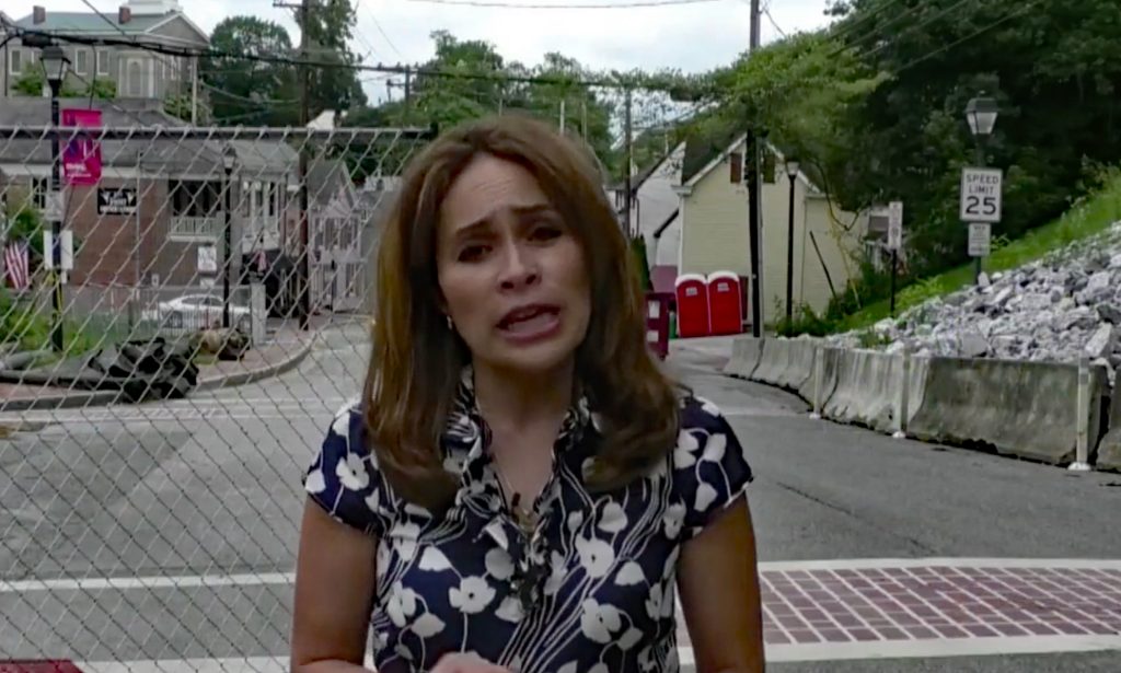 Female field reporter standing on a closed-off street in Ellicott City historic district, reporting on flood damage and recovery efforts after severe flooding.